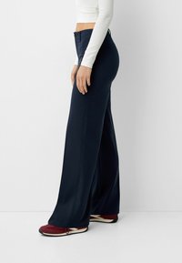 Navy wide-leg pants made of smooth fabric, featuring a high waist, front buttons, and side pockets, paired with red and cream sneakers.