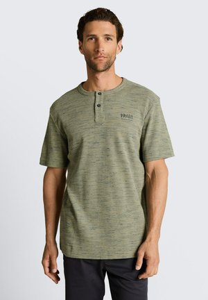 Mężczyzna o krótkich kręconych włosach, ubrany w oliwkowozieloną, teksturowaną koszulę henley z krótkim rękawem z dwoma guzikami i czarne spodnie, stojący na jednolitym tle.