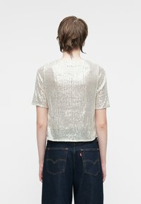 Top corto de manga corta con lentejuelas plateadas en líneas verticales, con un cuello redondo y un corte holgado. Combinado con pantalones de mezclilla oscuros.