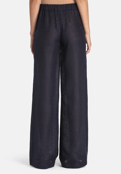 Vrouw die loszittende marineblauwe linnen broek met hoge taille en elastische tailleband draagt, gezien van achteren tegen een witte achtergrond.