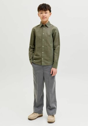 Dječak stoji noseći maslinasto zelenu košulju s gumbima, sive hlače i bež cipele, na jednostavnoj svijetloj pozadini.