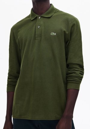 Tummaihoinen henkilö, joka käyttää oliivinvihreää, pitkähihakaista poolopaitaa, jossa on pieni kirjailtu krokotiililogo rinnassa.