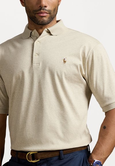 Tricou polo bej cu mâneci scurte și o închidere cu trei butoane, având un mic logo brodat pe partea stângă a pieptului. Purtați cu o curea maro.