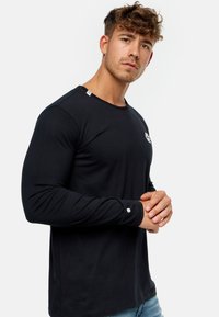 Camiseta negra de manga larga con cuello redondo, que presenta un logo blanco en el pecho y un pequeño emblema en la manga. Textura de algodón suave.
