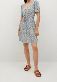 Blumenkleid mit V-Ausschnitt, kurzen Ärmeln und tailliertem Schnitt, in Blau mit orangefarbenen und gelben Mustern. Kombiniert mit braunen Riemensandalen.