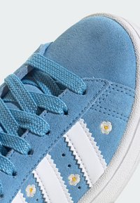 Sneaker in suede azzurro chiaro con strisce in pelle bianca, superficie testurizzata, dettaglio ricamato di margherita e lacci piatti blu. Suola in gomma visibile.