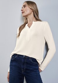 Cremfarbener Langarmtop mit Henley-Ausschnitt, kombiniert mit dunkelblauen Jeans. Weicher Stoff, lockere Passform und minimale Nähdetails.