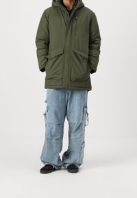 Vihreä eristetty parkatakki, jossa vetoketju edessä, huppu ja useita taskuja; yhdistetty väljälahkeisiin, vaaleansinisiin cargo-housuihin ja mustiin kenkiin.