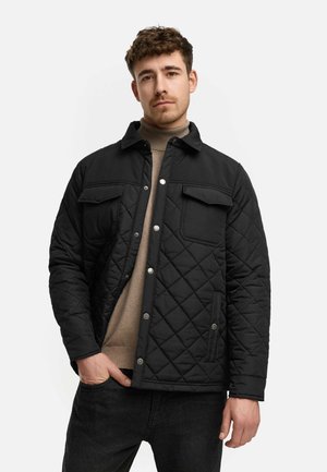 Svart quiltet jakke med krage, trykknapper og to frontlommer. Båret over en beige genser med glatt tekstur og moderne design.