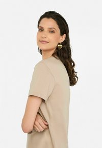 T-shirt beige à manches courtes, texture lisse, avec un col rond. Des boucles d'oreilles en forme de cœur doré ajoutent une touche de détail, le tout sur un fond neutre.