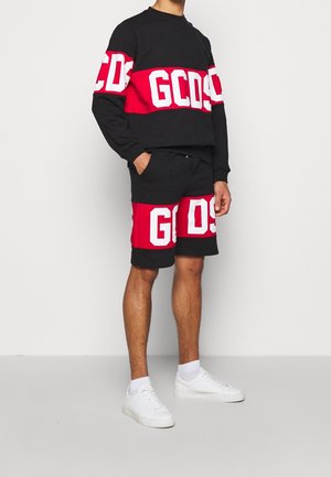 Homme portant un sweat-shirt et un short GCDS noirs et rouges avec des lettres blanches, assortis à des baskets blanches et des chaussettes, debout avec une main dans la poche.