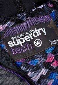 Sort stofmærke med "superdry" og "tech" i hvidt, med lilla accenter. Baggrunden viser et geometrisk mønster i pink og blå.