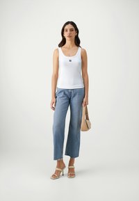 White sleeveless tank top with a small black logo, paired with light blue wide-leg jeans and beige high-heeled sandals. Holding a beige bag.