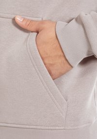 Lichtgrijs sweatshirtstof met een zachte textuur. Voorzien van een zijzak. Zichtbare geribde manchetten. Duim in de zak gestoken.