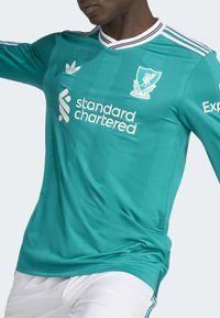 Maillot de football en bleu canard, avec des accents blancs, un col en V avec une bordure noire et des rayures horizontales. Les logos incluent Liverpool et Standard Chartered.