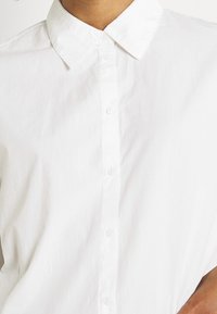 Bílá bavlněná košile s klasickým límečkem, která má knoflíkovou légou a hladkou, mírně zmačkanou strukturu. Design je minimalistický.