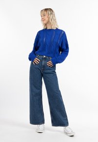 Pulover albastru tricotat cu modele din cablu, design scurt; purtat cu blugi largi, cu talie înaltă, de culoare albastră și adidași albi.