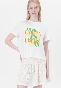 Fehér pamut póló, színes grafikai szöveggel és levél mintával, fehér hímzett szoknyával párosítva, amely texturált mintával rendelkezik.