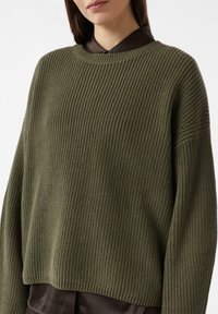 Khaki-grüner, gestrickter Pullover mit einer gerippten Struktur, überschnittenen Schultern und lockerer Passform, getragen über einem braunen Hemd mit Kragen.
