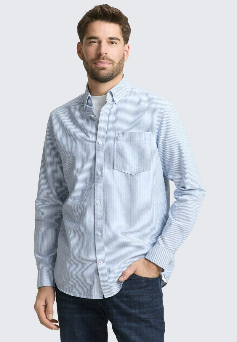 Uomo con barba che indossa una camicia a righe azzurre chiara con bottoni e jeans scuri, in piedi con una mano in tasca davanti a uno sfondo semplice.