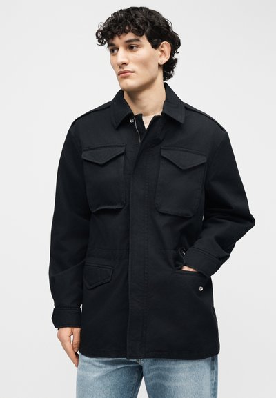 Νεαρός άνδρας με κατσαρά μαλλιά που φοράει μαύρο μπουφάν με τέσσερις τσέπες με καπάκι, με το ένα χέρι στην τσέπη, κοιτάζοντας προς τα πλάγια μπροστά σε απλό φόντο.