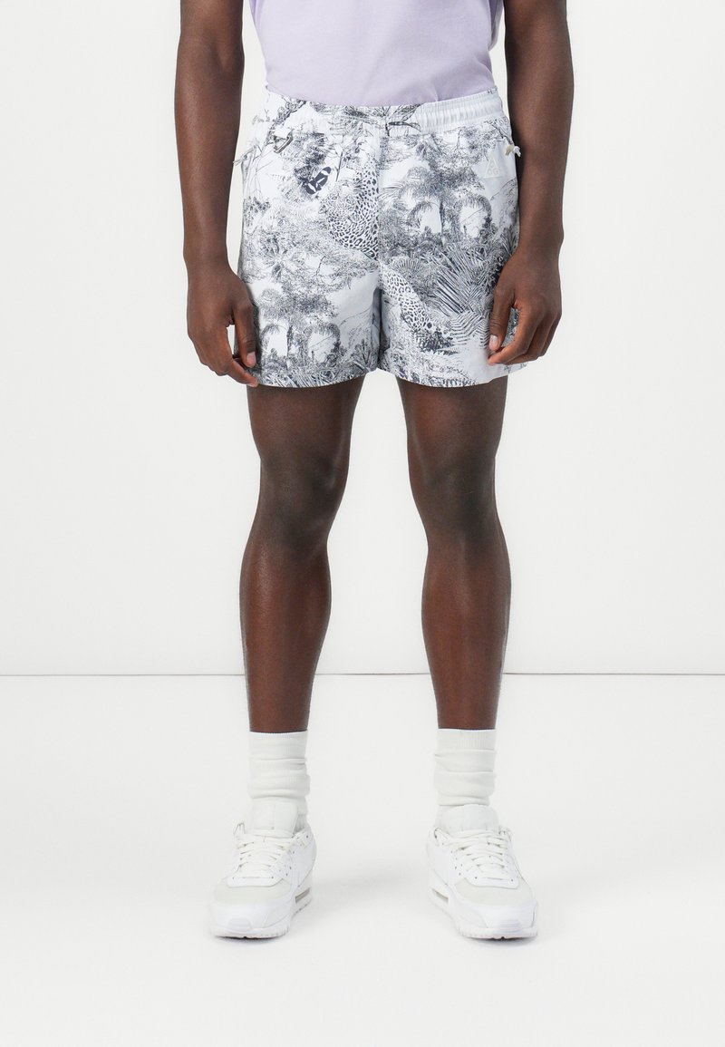 Swim shorts with a gray and black tropical pattern, elastic waistband, and side pockets, paired with white sneakers and crew socks.