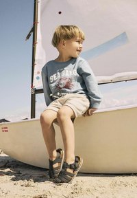 Brune sandaler med korksåler og mørke stropper; barn i blå sweatshirt med krabbe-tryk, beige shorts, siddende på en sejlbåd ved stranden.