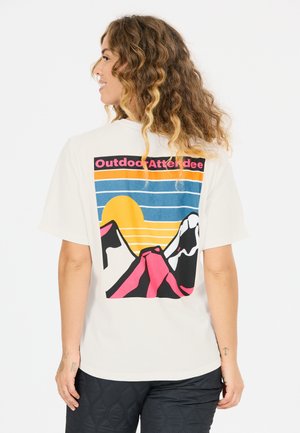 Frau mit lockigem Haar, die ein weißes T-Shirt trägt, auf dem auf der Rückseite eine bunte Berggrafik und der Schriftzug "Outdoor Attendee" abgebildet sind.