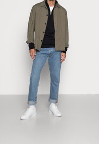 Veste vert olive, pull noir, jean bleu clair et baskets blanches. La veste est dotée de fermetures à boutons et d'une coupe décontractée.