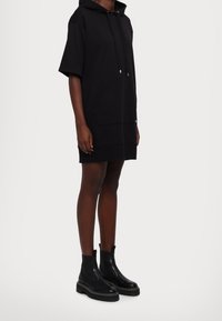 Czarna sukienka z kapturem, z krótkimi rękawami i przednią kieszenią. Posiada kaptur i boczne rozcięcie. Zestawiona z czarnymi botkami za kostkę na masywnych podeszwach.