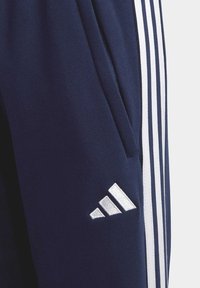 Navyblauwe joggingsbroek van zachte stof, met drie witte verticale strepen langs de zijkanten en een reflecterend zilveren Adidas-logo.