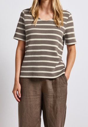 Vrouw draagt een kortärmelig top met bruine en witte strepen van textuurrijke stof en losse bruine broek, met één hand in de zak.