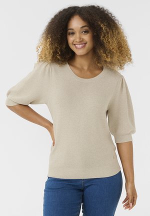 Mosolygó nő dús, göndör hajjal, bézs rövid ujjú pulóvert és kék farmert visel, egyik kezét a csípőjén tartva áll.