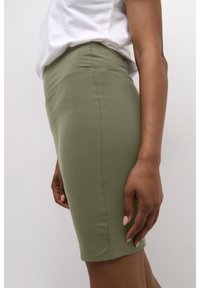 Femme portant une jupe ajustée vert olive et un haut blanc, montrée de la taille à la mi-cuisse, debout de profil avec le bras détendu.