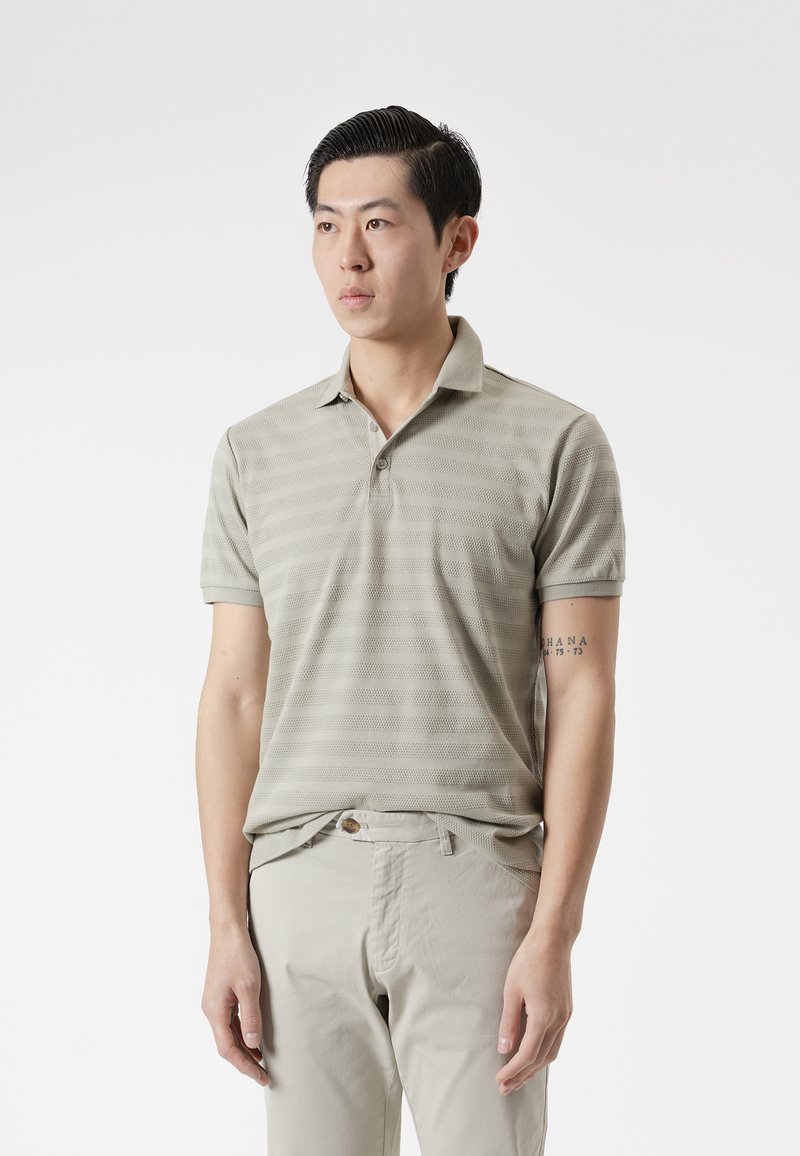 Hombre de cabello corto y negro, vistiendo una camiseta polo a rayas beige claro y pantalones beige, de pie con los brazos relajados contra un fondo blanco.