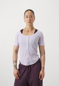 Haut court en ribbed lavande clair à manches courtes avec un col rond et des boutons sur le devant, superposé à un haut marron foncé ; associé à un pantalon violet ample.