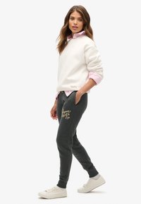 Biała bluza z okrągłym dekoltem i złotym logo; różowa koszula z kołnierzykiem pod spód; ciemnoszare dresy z złotym nadrukiem; białe sneakersy.