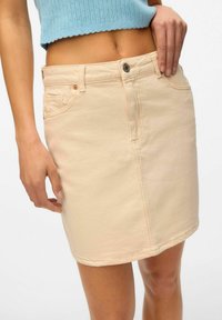 Minifalda de mezclilla beige con cierre de botón frontal, lazos para cinturón y bolsillos laterales. La tela tiene una textura suave y detalles de costura sutiles.