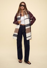 Veste en cuir bordeaux, crop top blanc, jean large bleu foncé, écharpe à carreaux marron et gris, et sandales sombres.