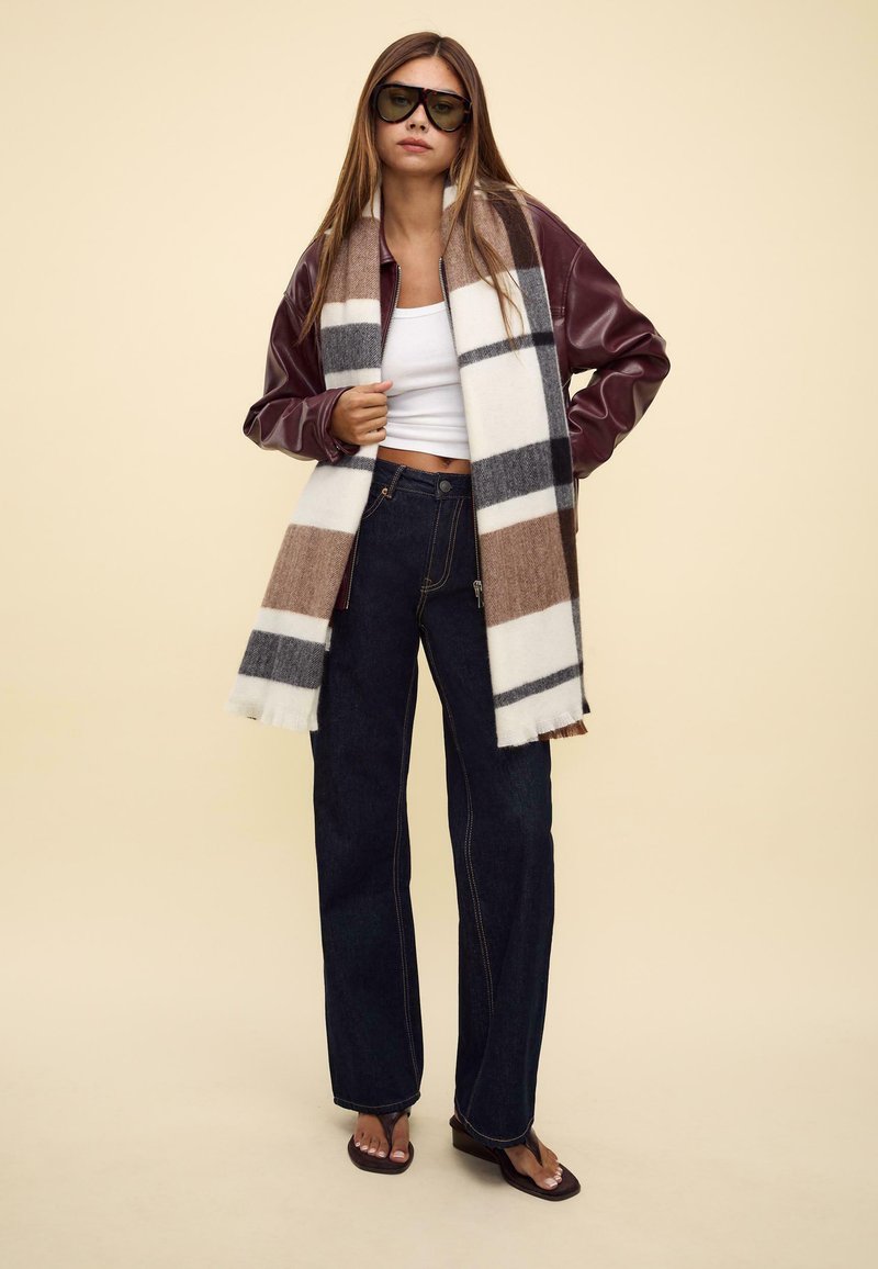 Veste en cuir bordeaux, crop top blanc, jean large bleu foncé, écharpe à carreaux marron et gris, et sandales sombres.