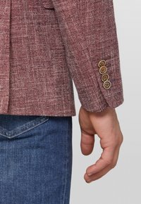 Blazer rosso con texture e un motivo sottile, con quattro bottoni marroni sui polsini. Abbinato a jeans in denim blu.