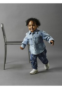 Camisa de mezclilla con dos bolsillos en el pecho, pantalones de chándal azul con un diseño de bicicletas y zapatillas blancas. Niño de pie cerca de una silla.