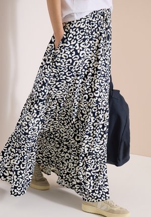 Persona che indossa una lunga gonna a stampa leopardata bianca e nera, sneakers bianche e tiene una borsa blu scuro, in piedi su uno sfondo chiaro.