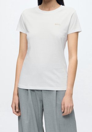 Femme portant un t-shirt blanc uni à manches courtes avec un petit logo "BOSS" sur la poitrine et un pantalon plissé gris clair.