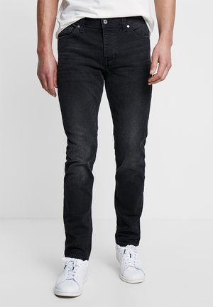 Mand iført slim-fit sorte jeans og hvide sneakers stående mod en ensfarvet lys baggrund.