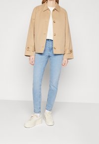 Beige bavlněná bunda s cropped střihem, velkými knoflíky a ohrnutými manžetami, v kombinaci se světle modrými skinny džíny a krémovými teniskami.