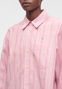 Gros plan d'une personne portant une chemise rose à boutons avec de fines rayures verticales et une poche poitrine à l'avant.