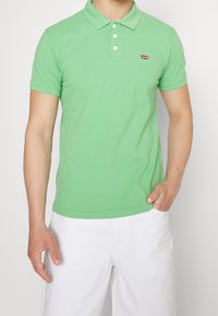 Man som bär en ljusgrön kortärmad pikétröja med vita knappar och Levi's-logotyp, tillsammans med vita shorts, med handen i fickan.