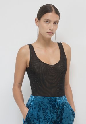 Donna con capelli tirati indietro, che indossa lunghi orecchini a catena, un top in rete nero con borchie e pantaloni di velluto blu, con le mani in tasca.