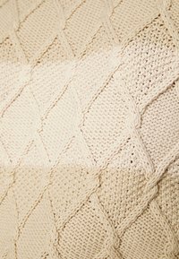 Coperta lavorata a maglia color beige, con un motivo a diamante testurizzato che presenta diverse tecniche di lavorazione e transizioni di colore morbide.